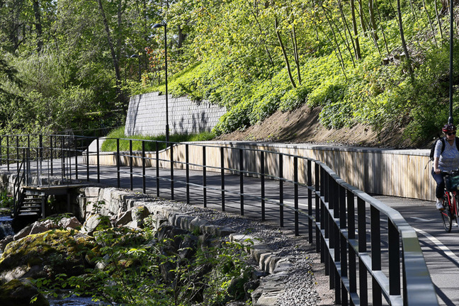 En person cyklar på en cykelbana bredvid ett vattendrag med stenar och grönska i skogsmiljö.